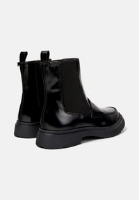 Botas Chelsea negras y brillantes con paneles elásticos en los laterales, que presentan una suela gruesa negra y tiradores en la parte posterior para ponerlas y quitarlas fácilmente.