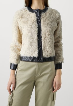 Woman wearing a cream textured fur jacket with black leather trim and beige utility pants, holding a small round silver object in left hand.