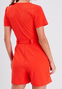 Combinaison courte à manches courtes rouge en tissu extensible, dotée d'une taille ajustée avec ceinture et d'une texture lisse avec un short ajusté.