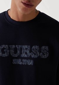 Felpa blu navy con collo rotondo, con un logo "GUESS" in rilievo con cuciture azzurre più chiare e "EST. 1981" sotto. Tessuto morbido.