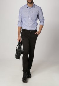 Blau-weißer gestreifter Button-Up-Hemd, schwarze Hose und schwarze Stiefel. Hält eine schwarze Tasche und steht vor einem neutralen Hintergrund.