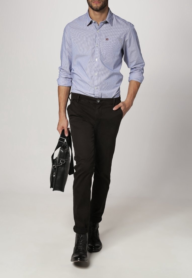 Blau-weißer gestreifter Button-Up-Hemd, schwarze Hose und schwarze Stiefel. Hält eine schwarze Tasche und steht vor einem neutralen Hintergrund.