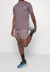 Hombre estirando los cuádriceps mientras lleva una camiseta púrpura, pantalones cortos a juego, calcetines negros y zapatillas deportivas azules, contra un fondo blanco.