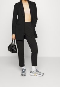 Svart skräddarsydd kavaj över en nude bralette, ihopkopplad med svarta byxor, vita strumpor och silvriga sportskor med blå accenter. Svart handväska ingår.