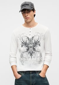 Λευκό πουκάμισο με μακριά μανίκια και υφή φινιρίσματος, με μαύρο γραφικό σχέδιο που περιλαμβάνει στιλιζαρισμένα κέρατα και κείμενο. Συνδυάζεται με γκρι καπέλο.