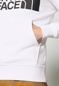 Mano parzialmente dentro della tasca anteriore di una felpa bianca con logo nero, indossata sopra pantaloni marroni su uno sfondo chiaro.