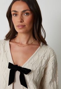 Crème gebreide cardigan met een kabelpatroon, met een zwarte fluwelen strik bij de halslijn. Model draagt een eenvoudig topje eronder.