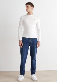 Vit långärmad skjorta, blå denimjeans och vita sneakers. Skjortan har rundad halsringning och en kroppsnära design; jeansen är rakskurna med grundläggande fickor.