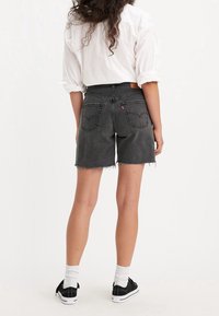 Shorts de denim negro con dobladillo deshilachado, cintura alta y dos bolsillos traseros, combinados con una camisa blanca y zapatillas negras.