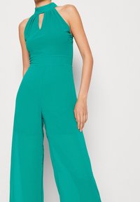 Mono halter en azul verde fabricado con tejido suave, que presenta un detalle de ojal en el escote y un diseño de pierna ancha, creando una silueta fluida.