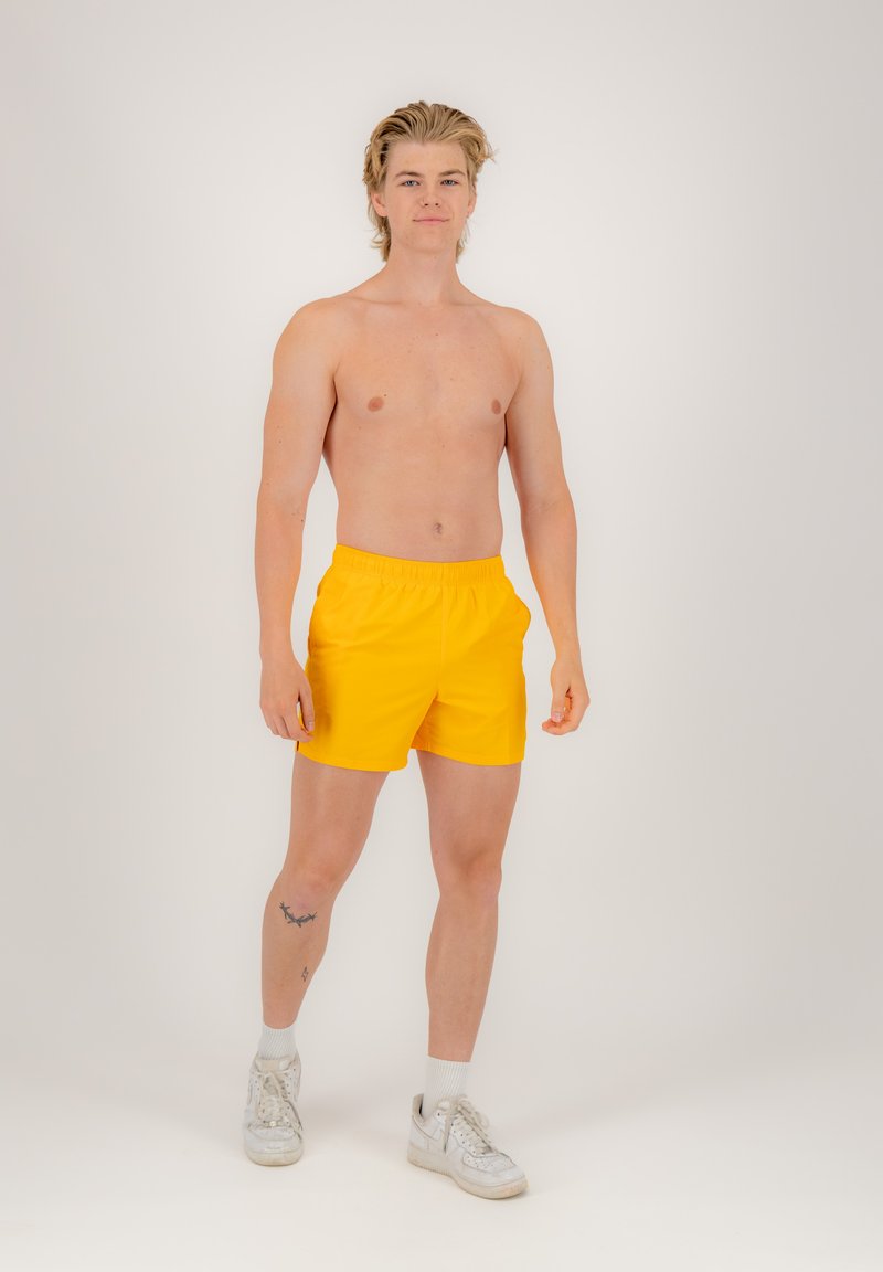 Żółte szorty kąpielowe z elastycznym paskiem w talii, wykonane z gładkiego, lekkiego materiału; model nosi białe trampki i skarpetki sięgające kostek.