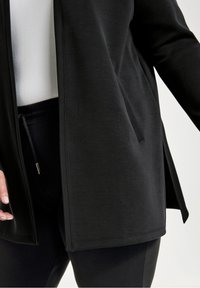 Černá bunda s dlouhým rukávem z měkké látky, s rovný střihem, otevřeným předním dílem a bočními kapsami, doplněná černými kalhotami.