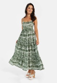 Green and white patterned maxi dress with spaghetti straps, tiered design, and a flowy silhouette; paired with white, chunky-heeled sandals.
