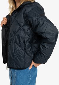 Veste matelassée noire avec un design matelassé, dotée d'une capuche, d'une fermeture éclair et de poignets côtelés. Le matériau a une texture brillante, léger.