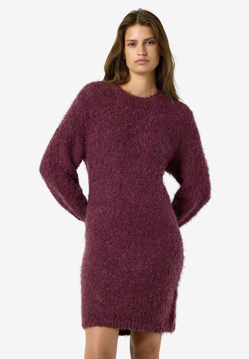 Femme portant une robe-pull en maille violette foncée et duveteuse à manches longues, debout les mains derrière le dos sur un fond blanc uni.