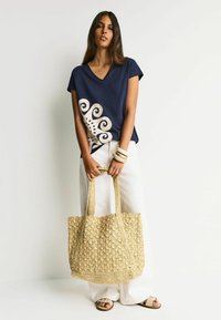 Granatowa bluzka z dekoltem w serek, z kremowym spiralnym wzorem po jednej stronie, zestawiona z białymi spodniami o szerokich nogawkach. Trzyma plecioną słomianą torbę typu tote z fakturowanymi detalami.