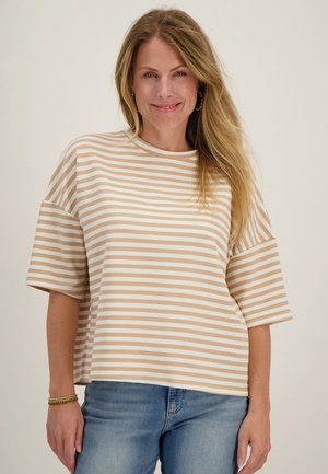 Vrouw met lang blond haar, die een beige-wit gestreept oversized shirt met korte mouwen en een blauwe spijkerbroek draagt, glimlacht tegen een effen achtergrond.