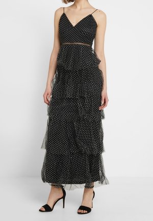 Sort ærmeløs maxi-kjole med hvide polkaprikker, lagdelt tylskørt, tynde stropper og et mesh-detalje i taljen, båret med sorte højhælede sandaler.
