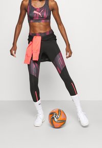 Černé sportovní legíny s růžovými akcenty a vzorem, doplněné sladěným sportovním topem. Růžová mikina uvázaná v pase a fotbalový míč.