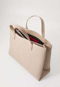 Beige skinnveske med en teksturert overflate, to håndtak og en glidelås lomme. Har en merket logo på fronten, og interiøret har røde detaljer.