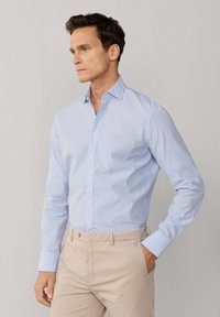 Homme portant une chemise habillée à carreaux bleu clair rentrée dans un pantalon beige, debout avec une main dans la poche contre un fond uni.