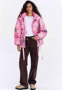 Różowa pikowana kurtka z kwiatowym wzorem, z wysokim kołnierzem i sznurkami ściągającymi. W zestawie z brązowymi spodniami o szerokich nogawkach i jasnymi butami.