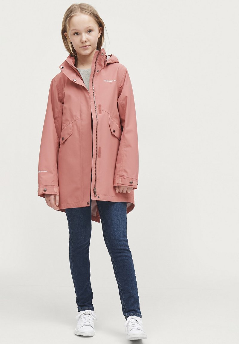 Waterdichte roze parka jas met capuchon, rits aan de voorkant en twee zijzakken, gecombineerd met donkerblauwe jeans en witte sneakers.