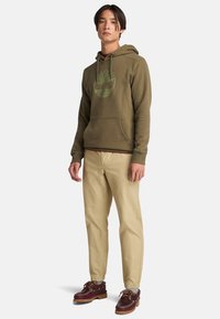 Felpa verde oliva con tasca a canguro frontale e logo ad albero, abbinata a pantaloni beige e scarpe marroni con dettagli gialli e lacci.