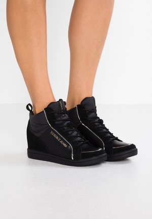 Baskets compensées noires avec lacets et logo "Versace Jeans" portées aux pieds sur fond blanc.