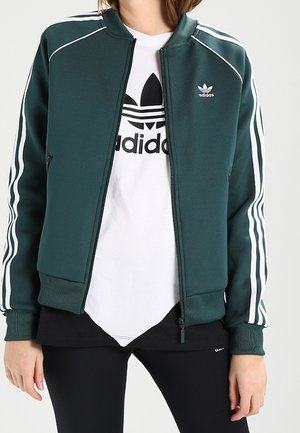 Persoon draagt een donkergroene Adidas zip-up jas met witte strepen op de mouwen over een wit T-shirt met Adidas-logo en zwarte legging.
