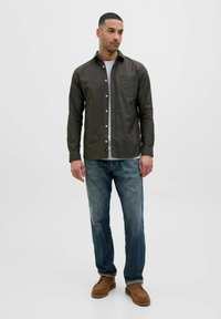 Camicia button-up verde oliva scuro con taschino, bottoni bianchi, indossata sopra una maglietta grigia, abbinata a jeans blu e scarpe marroni.
