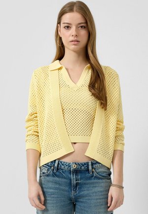 Jeune femme aux longs cheveux portant un cardigan en maille jaune et un top court assorti avec un jean bleu, regardant vers l'avant sur un fond uni.