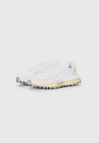 Zapatillas Lacoste con parte superior de tela blanca y detalles de ante, entresuela beige y suela de goma gris con picos de tracción.