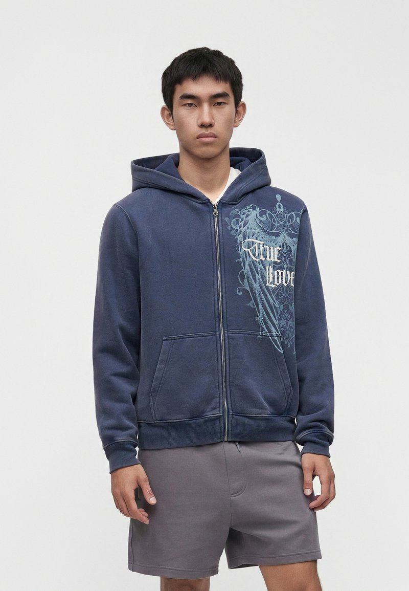 Jeune homme portant un sweat à capuche zippé bleu marine avec le texte « True Love » et un dessin d'ailes sur la poitrine, associé à un short gris, debout devant un fond uni.