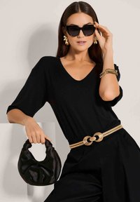 Zwarte v-hals top, gecombineerd met een riem met gouden accenten en cirkelvormige hardware. Accessoires zijn onder andere grote zonnebrillen en een zwarte schoudertas.