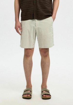 Pantalones cortos a rayas en tela beige claro y blanca, con un corte relajado. Combinados con sandalias verdes tipo deslizador. La blusa es un tejido marrón texturizado.