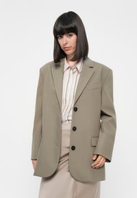 Khakifarbenes Oversized-Blazer mit strukturierten Schultern, einer einzelnen Brusttasche und drei schwarzen Knöpfen, kombiniert mit einem gestreiften Hemd.