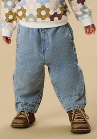 Svetlo modre široke jeans hlače z elastičnim pasom in stranskimi žepi, kombinirane s rjavimi škornji z vezalkami. Ozadje je enobarvno bež.