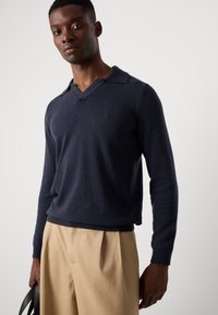 Navy blue knitted sweater with a V-neck and long sleeves, featuring a subtle logo on the chest and a relaxed fit paired with beige trousers.