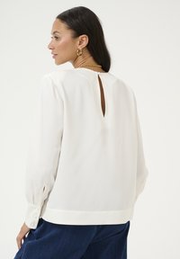 Witte blouse met lange mouwen en een gestructureerde afwerking, met een keyhole rugdetail en met knopen gesloten manchetten, gecombineerd met donkere spijkerbroek.