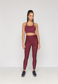 Kobieta stoi w bordowym staniku sportowym i dopasowanych legginsach wysokotaliowych, nosząc czarne i białe buty sportowe, na neutralnym tle.