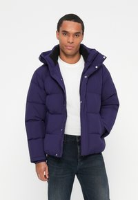 Man wearing a dark blue puffer jacket over a white shirt and dark jeans, standing against a plain white background.