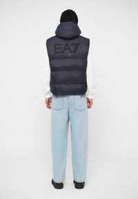 Jūras spēku puffera vests ar kapuci, ar quiltota dizaina daļām un izšūtu "EA7" logo aizmugurē, saskaņots ar gaiši ziliem džinsiem.