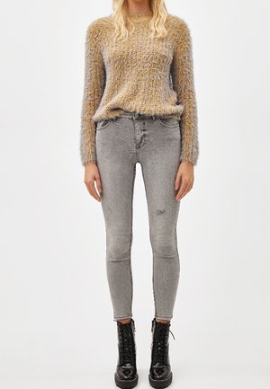 Femme portant un pull en maille jaune duveteux, un jean skinny gris et des bottines noires à lacets, debout devant un fond blanc uni.