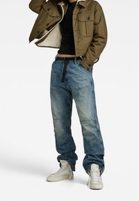 Olivgrön kort jacka med fårskinnsfoder, kombinerad med slitna blåjeans med lappar. Vita hög-topp sneakers.