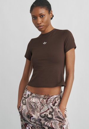 Femme portant une chemise brune cintrée à manches courtes avec un petit logo blanc, pantalon à motif cachemire, les mains dans les poches sur un fond uni.