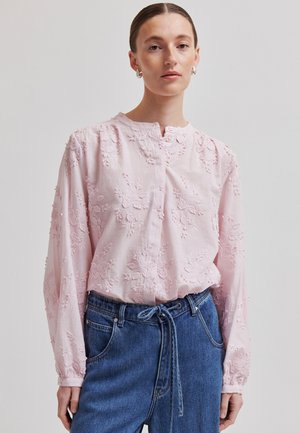 Frau trägt eine blassrosa, floral bestickte Bluse mit langen Ärmeln und hoch taillierte blaue Jeans, die mit einem Jeansgürtel gebunden sind.