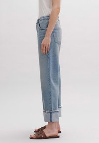 Vaqueros de mezclilla azul claro de pierna recta con dobladillo deshilachado y puños enrollados. Combinados con sandalias de dedo marrones. Diseño minimalista, sin adornos.