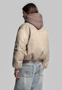 Beige pufferjakke med ribbede manchetter og talje, prydet med en brun hætte og rygbroderi. Kombinationen af let denimbukser med logo.