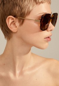 Occhiali da sole tartarugati marroni con una forma quadrata audace, accentuati in oro sulle tempie e con lenti scure, indossati con capelli corti e trucco minimale.
